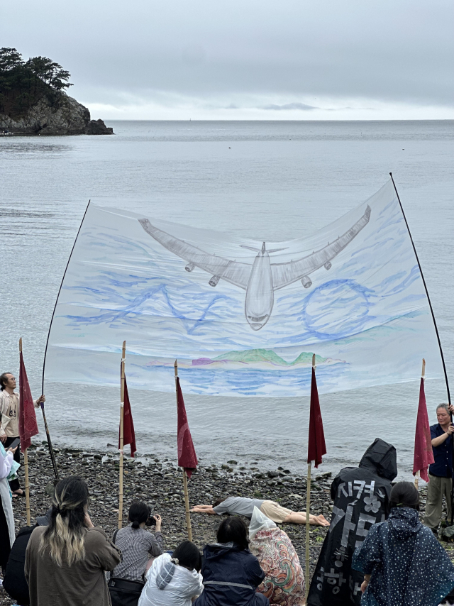 14일 오후 부산 강서구 가덕도 외양포 일대에서 열린 '가덕신공항반대 예술행동' 퍼포먼스 모습. 김화순 작가가 자신이 직접 그린 걸개그림 아래에 누워서 퍼포먼스를 펼치고 있다. 김은영 기자